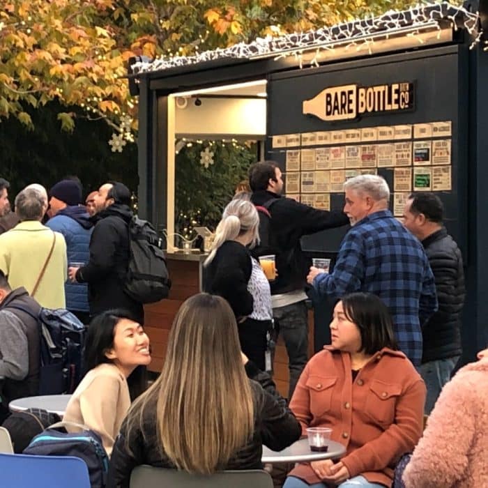 Bare Bottle outdoor walk-up window with people standing and sitting around front with a fall colored tree in the backgorund
