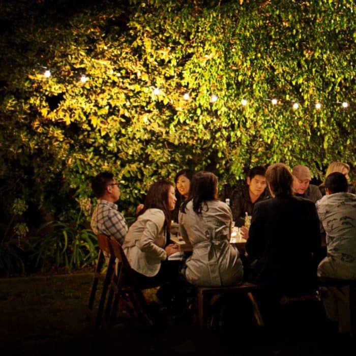 People conversing, sitting around an outdoor table in the evening with string lights above and foliage in the background enclosing the space