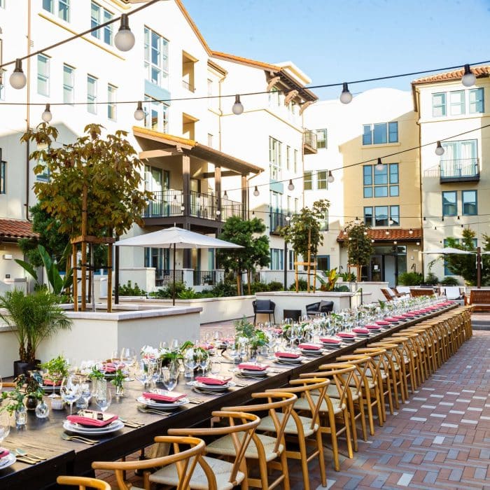 Elegant outdoor dining setup with long tables adorned with glassware and red napkins, surrounded by wicker chairs. Overhead string lights and modern apartment buildings in the background convey a festive, sophisticated ambiance.