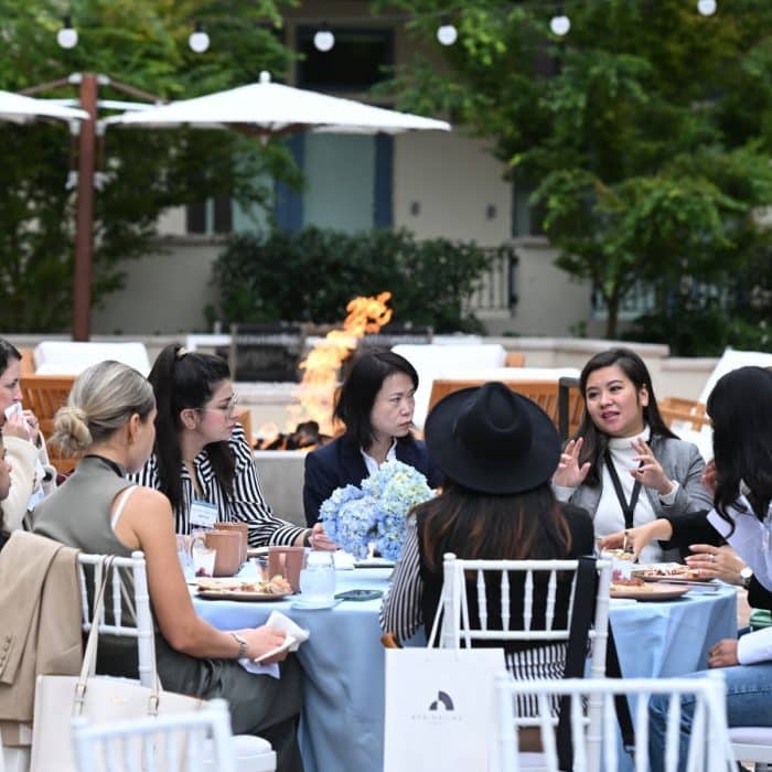 A group of seven women sit around a round table with a blue tablecloth, engaged in animated conversation. A fire pit glows warmly behind them.