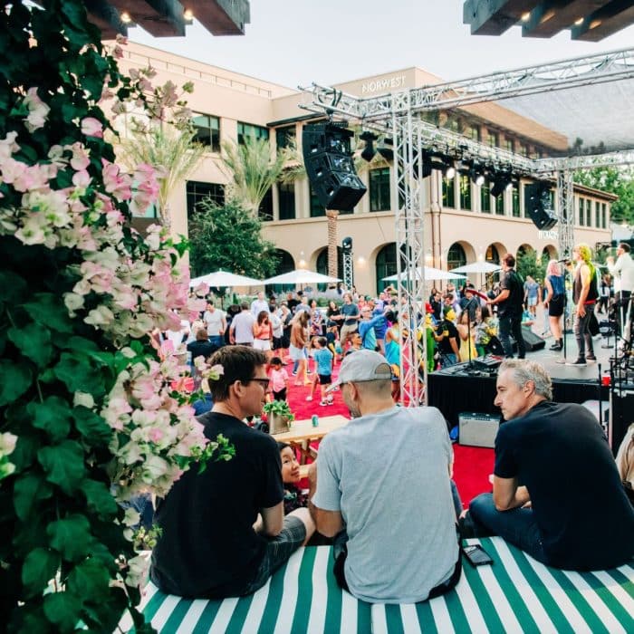Outdoor concert scene with a vibrant crowd, a band performing on stage, and three men sitting on a striped bench surrounded by blooming flowers.