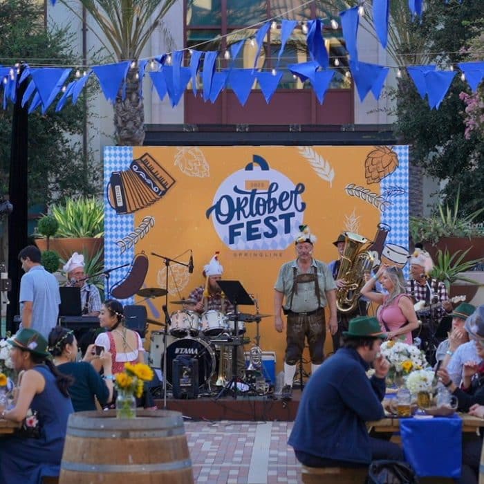 A festive Oktoberfest scene with a band performing beneath blue and white bunting. People in traditional Bavarian attire enjoy the lively atmosphere.