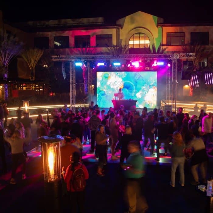 Outdoor night party with a lively crowd dancing under colorful lights. A DJ booth is in the background, projecting vibrant visuals. Atmosphere is energetic and festive.