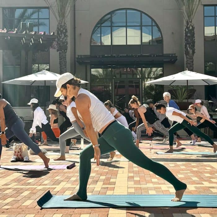 A group of people practice yoga outdoors on a sunny day. They stand in a bent posture on mats, surrounded by brick pavement and green plants.